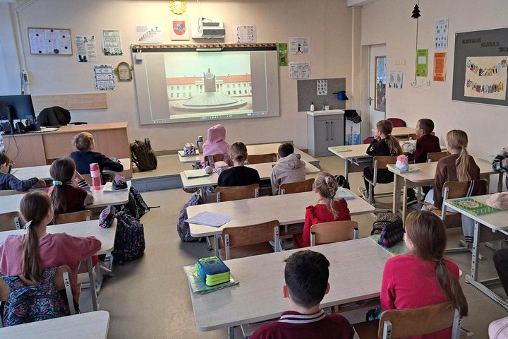 klasėje sėdintys trečios klasės mokiniai klausosi edukacijos apie Lietuvos valstybės sukūrimą; priekyje matomas projektorius su istoriniu vaizdu, vaikai sėdi suoluose ir žiūri į ekraną.