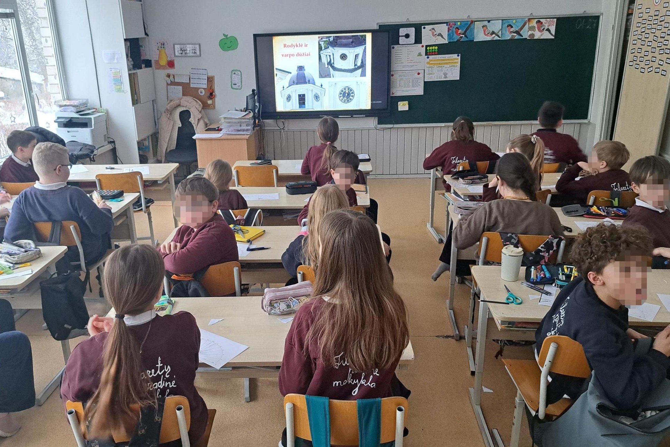 Klasėje sėdintys pradinių klasių mokiniai dalyvauja edukacijoje apie Vilniaus gimtadienį ir Vilniaus pilis, priekyje ekrane matoma pristatymo skaidrė su Vilniaus bokšto laikrodžiu.