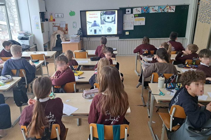 Klasėje sėdintys pradinių klasių mokiniai dalyvauja edukacijoje apie Vilniaus gimtadienį ir Vilniaus pilis, priekyje ekrane matoma pristatymo skaidrė su Vilniaus bokšto laikrodžiu.