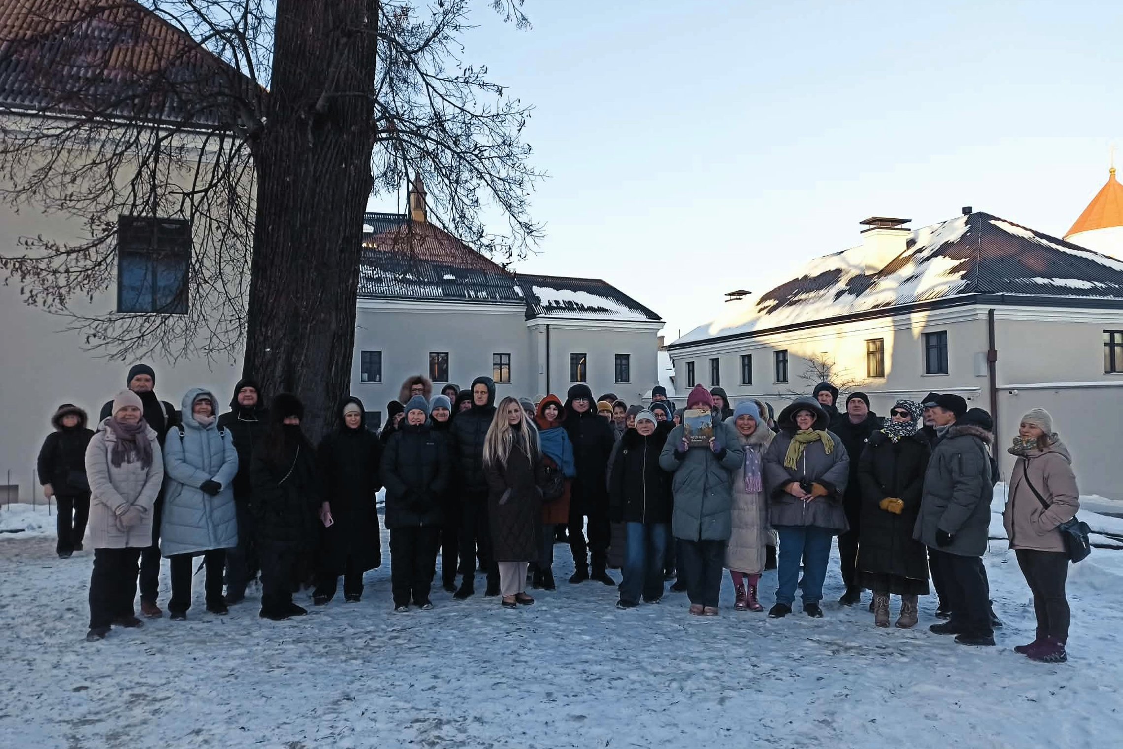 Ekskursijos dalyviai stovi Vilniaus pilių teritorijoje žiemą, tarp istorinių pastatų, apsirengę šiltais drabužiais.