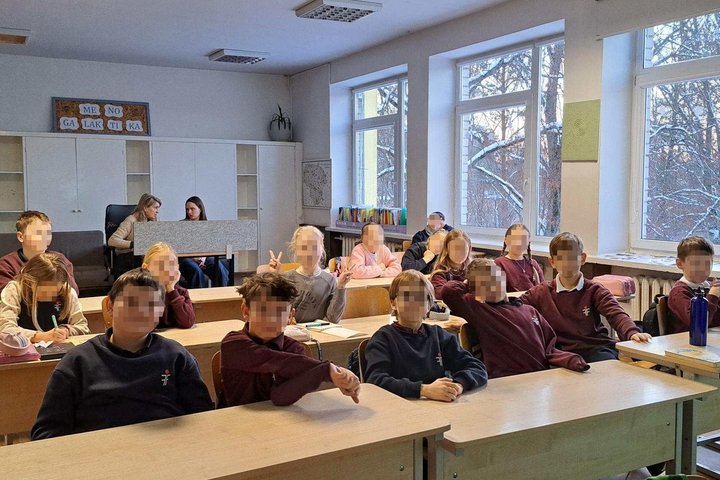 Klasėje prie suolų sėdi mokiniai, dalyvaujantys edukacijoje – vaikai klausosi, rašo ir aktyviai įsitraukia į užsiėmimą, pro didelius langus matyti žiemos vaizdas.