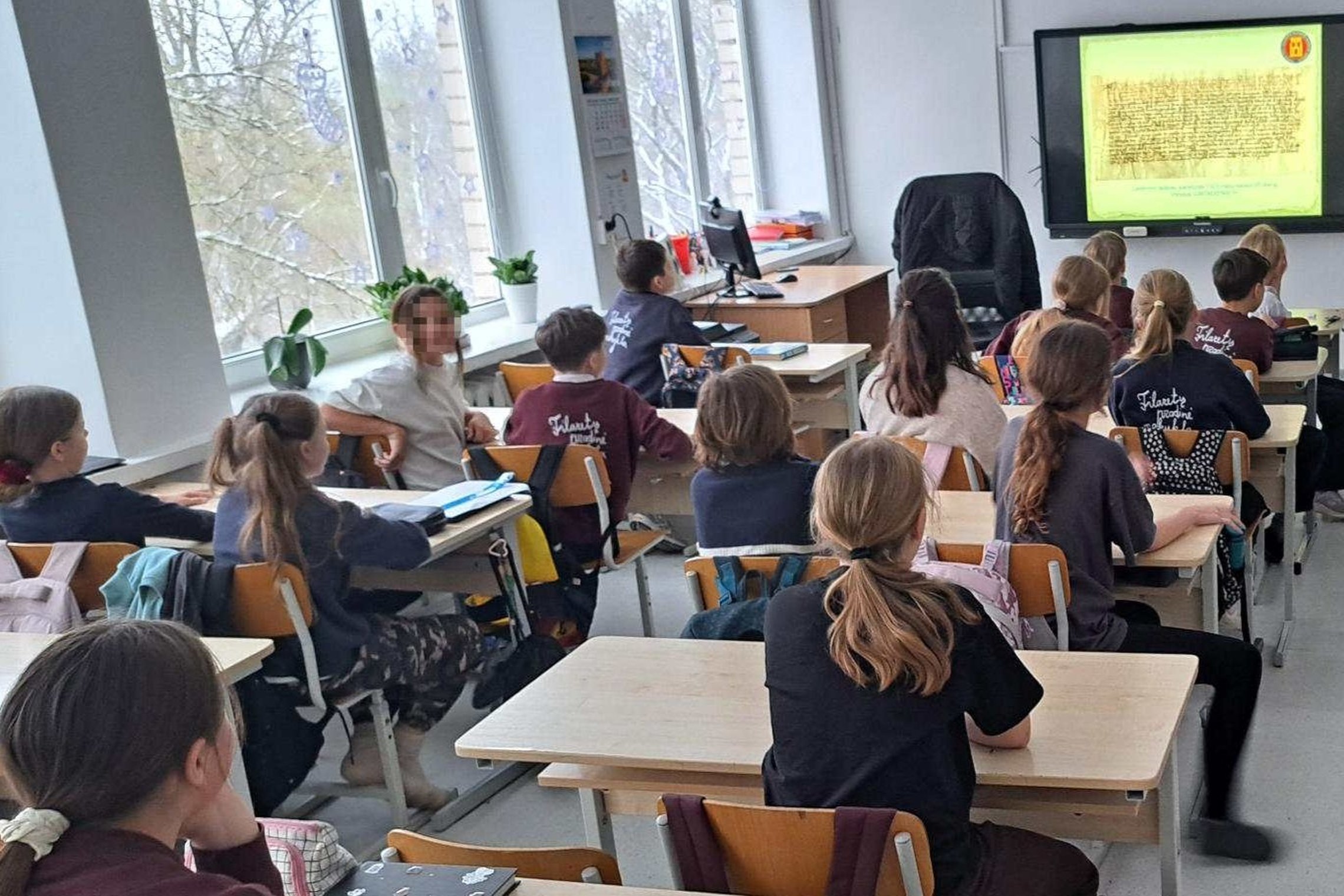 Vilniaus Filaretų gimnazijos ketvirtokai sėdi klasėje ir dalyvauja edukacijoje apie Didžiuosius Lietuvos kunigaikščius, klausydamiesi pristatymo ekrane.