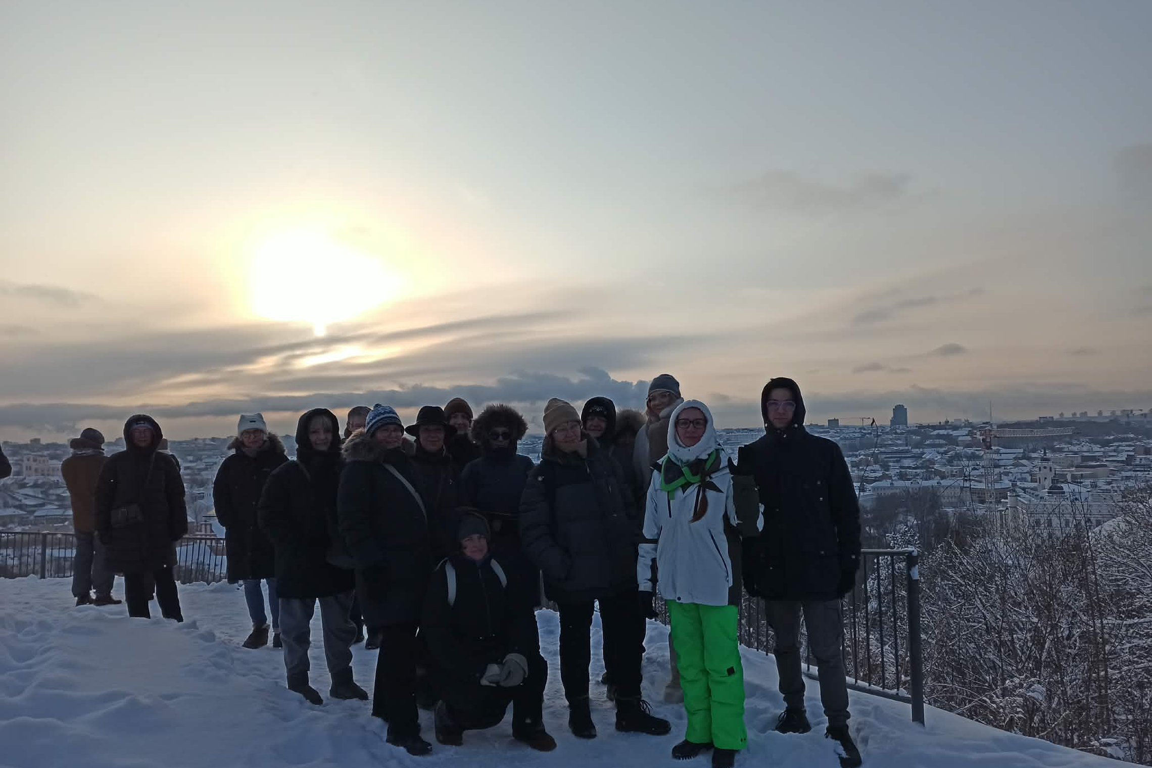 Grupė žmonių stovi ant apsnigtos apžvalgos aikštelės ir fotografuojasi žiemą. Už jų matyti snieguotas Vilniaus miestas, o danguje leidžiasi saulė