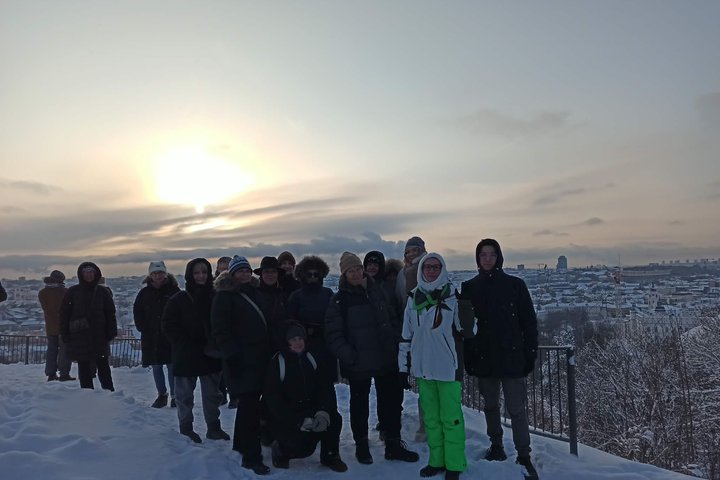 Grupė žmonių stovi ant apsnigtos apžvalgos aikštelės ir fotografuojasi žiemą. Už jų matyti snieguotas Vilniaus miestas, o danguje leidžiasi saulė