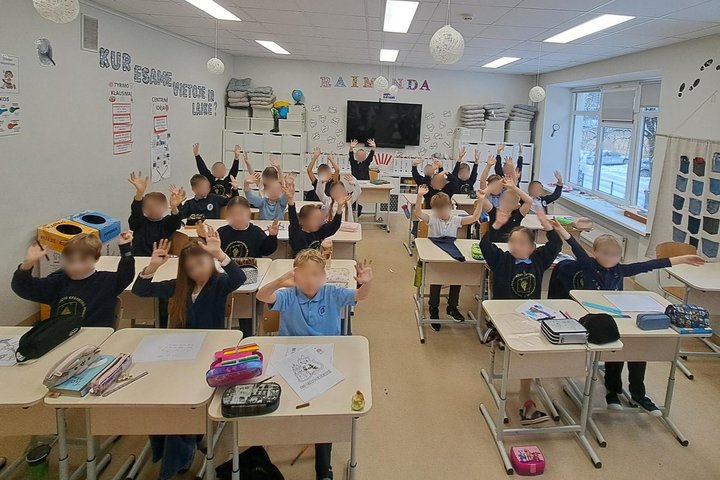 Nuotraukoje vaikai klasėje, sėdi suoluose ir mojuoja. Linksmi po edukacijos apie sostinės gimtadienį.