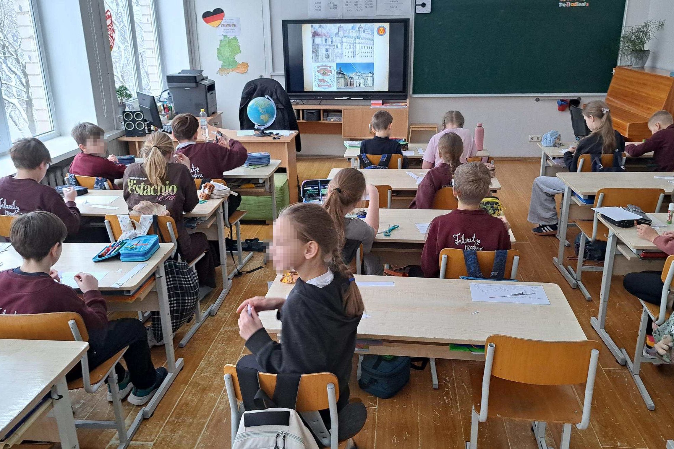 ALT: Klasėje sėdintys mokiniai prie suolų atlieka užduotis, o priekyje interaktyviame ekrane matomi pilies vaizdai; šalia mokytojo stalo stovi gaublys, už nugaros – lenta ir pianinas, pro langus matyti žiemos peizažas.