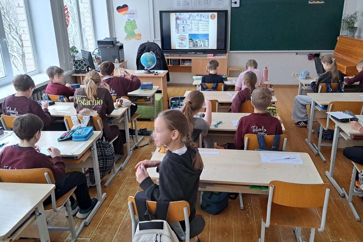 ALT: Klasėje sėdintys mokiniai prie suolų atlieka užduotis, o priekyje interaktyviame ekrane matomi pilies vaizdai; šalia mokytojo stalo stovi gaublys, už nugaros – lenta ir pianinas, pro langus matyti žiemos peizažas.