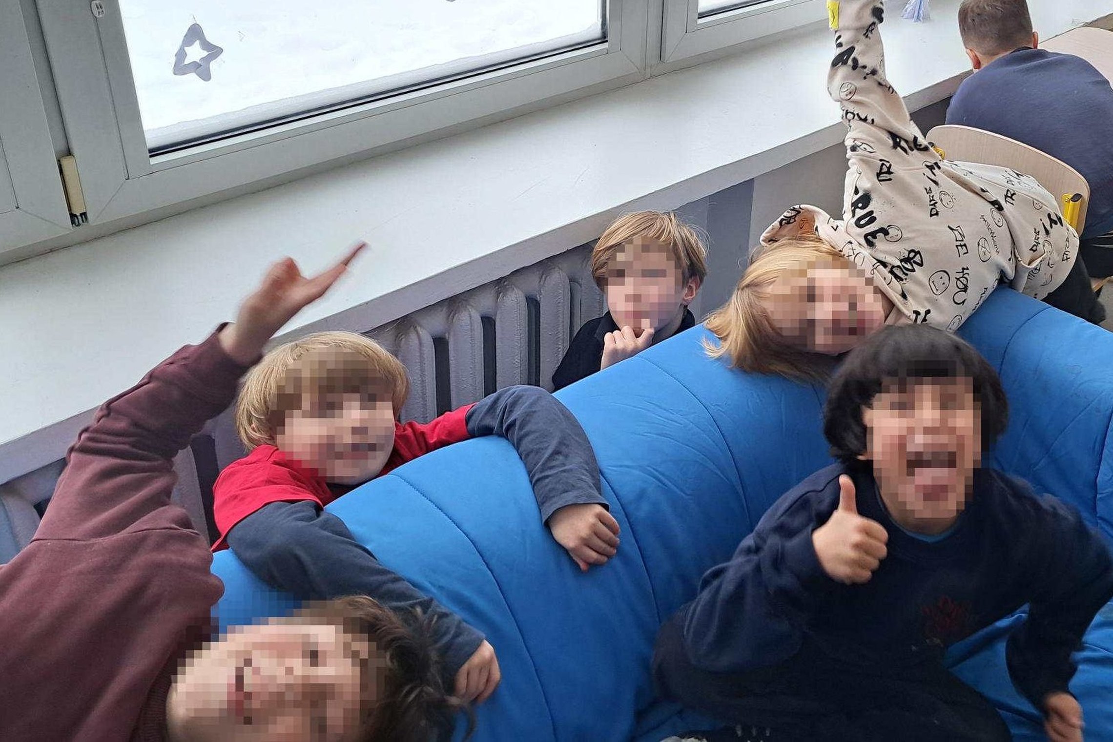 Pradinės mokyklos klasėje antrokai dalyvauja edukacijoje – vaikai sėdi ir žaidžia ant mėlyno minkšto baldo, keli pakėlę rankas ar rodantys nykštį į viršų, fone matyti mokykliniai suolai ir langai