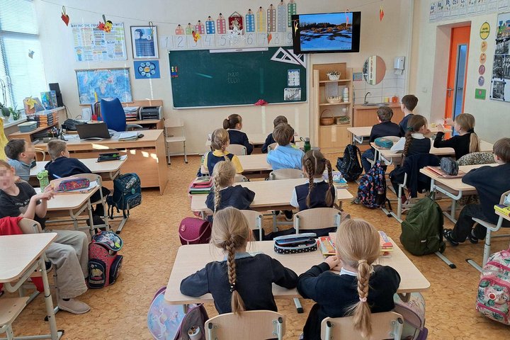 Pradinukai sėdi klasėje suole ir stebi ekrane rodomą vaizdą, vykstant edukaciniam užsiėmimui apie žmonių gyvenimą praeityje.