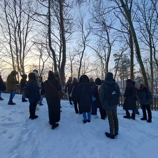Žmonių grupė stovi snieguotame miško take prie medinės tvoros ir klausosi gido. Aplink – aukšti be lapų medžiai, o pro jų šakas matyti žiemos vakaro dangus.