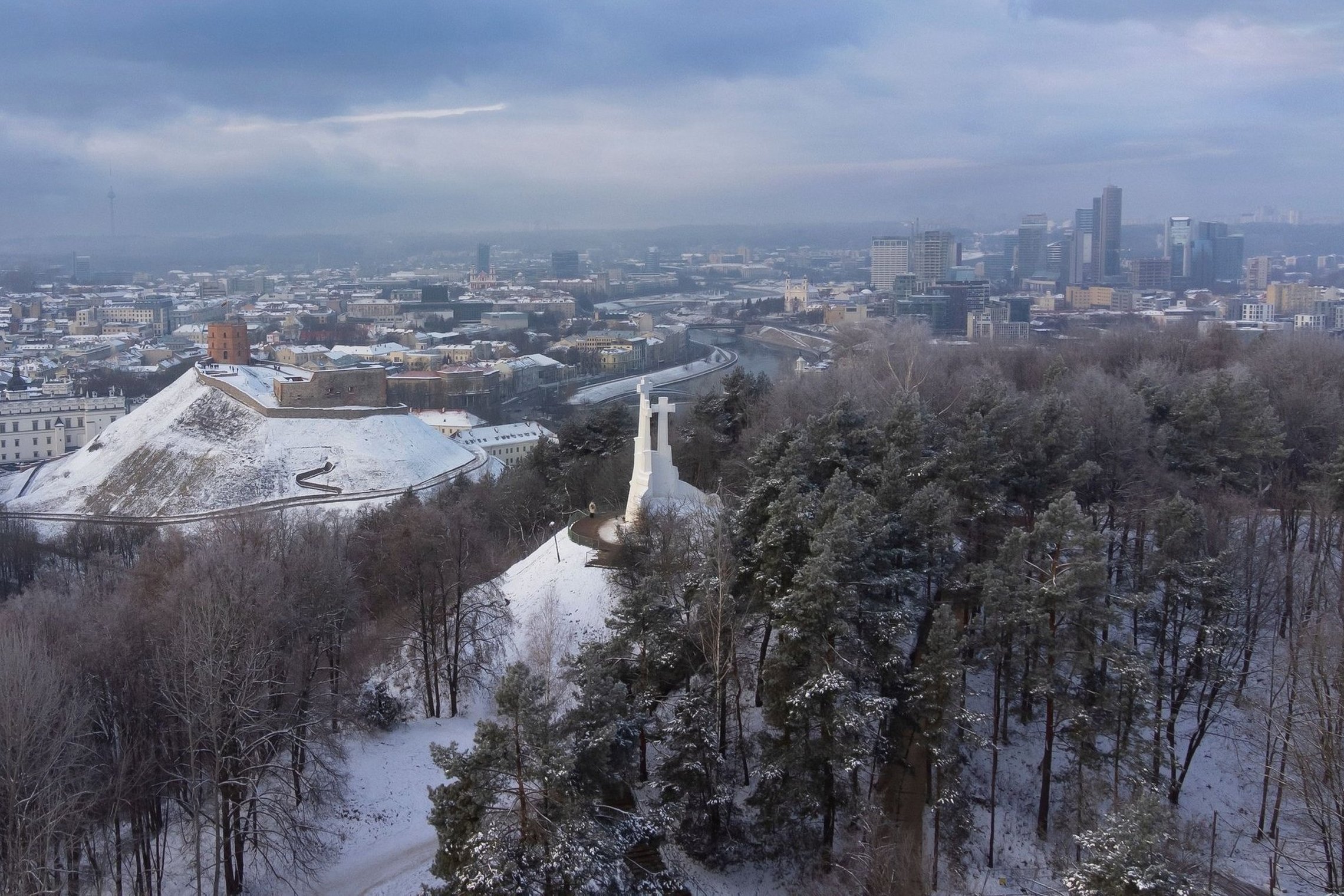 Žiemiška Vilniaus panorama iš viršaus: apsnigtas Gedimino kalnas su bokštu, Trijų Kryžių paminklas ant kalvos, aplink miškai ir miesto pastatai, tolumoje matyti modernių dangoraižių kvartalas po debesuotu dangumi