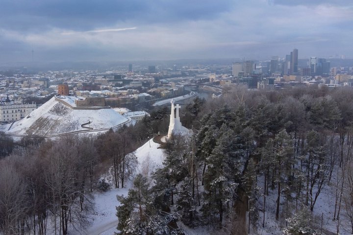 Žiemiška Vilniaus panorama iš viršaus: apsnigtas Gedimino kalnas su bokštu, Trijų Kryžių paminklas ant kalvos, aplink miškai ir miesto pastatai, tolumoje matyti modernių dangoraižių kvartalas po debesuotu dangumi