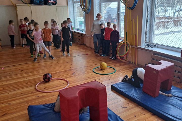 Pradinės mokyklos sporto salėje vaikai dalyvauja judrioje pamokoje – eina per kliūčių ruožą su lankais, kamuoliais ir čiužiniais, o mokytoja juos stebi ir lydi; pro langus matyti žiemos vaizdas lauke.
