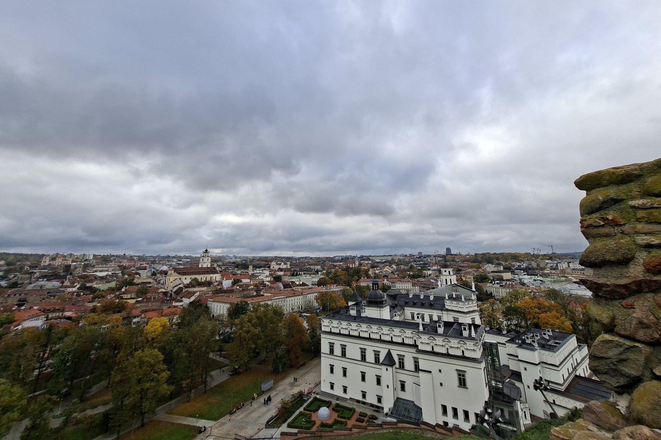 Panoraminis Vilniaus senamiesčio vaizdas nuo aukštos apžvalgos vietos. Nuotraukoje matyti balti Lietuvos nacionalinio muziejaus pastatai, aplink juos augantys rudens spalvomis nusidažę medžiai ir tankiai užstatyta miesto panorama. Dešinėje kadro pusėje ma