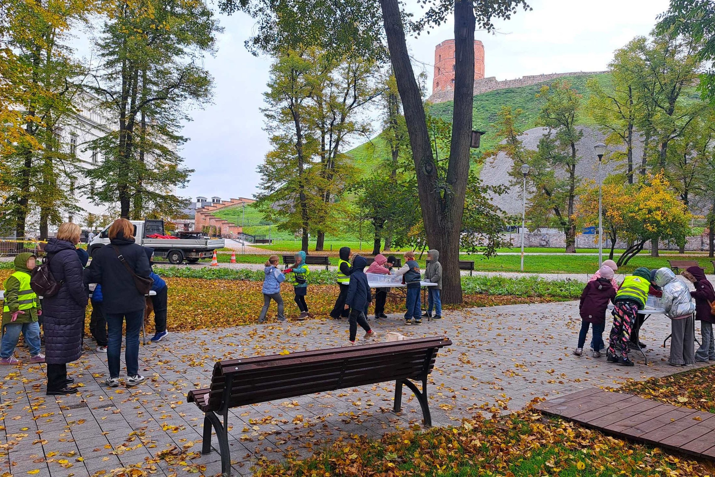 Vilniaus pilių teritorijoje, Gedimino pilies bokšto papėdėje, rudeniškame parke pradinukai dalyvauja edukacinėje veikloje prie lauke pastatytų stalų. Vaikai su ryškiomis liemenėmis atlieka užduotis, o juos supa spalvingi medžiai ir nukritę rudens lapai.