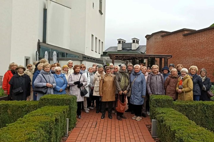 Dalyviai ekskursijoje „Vyšnios kaip apelsinai: Vilniaus pilių sodai ir parkai“. Senjorai pozuoja bendrai nuotraukai lauke, tarp žalių gyvatvorių, istorinių pastatų apsuptyje.