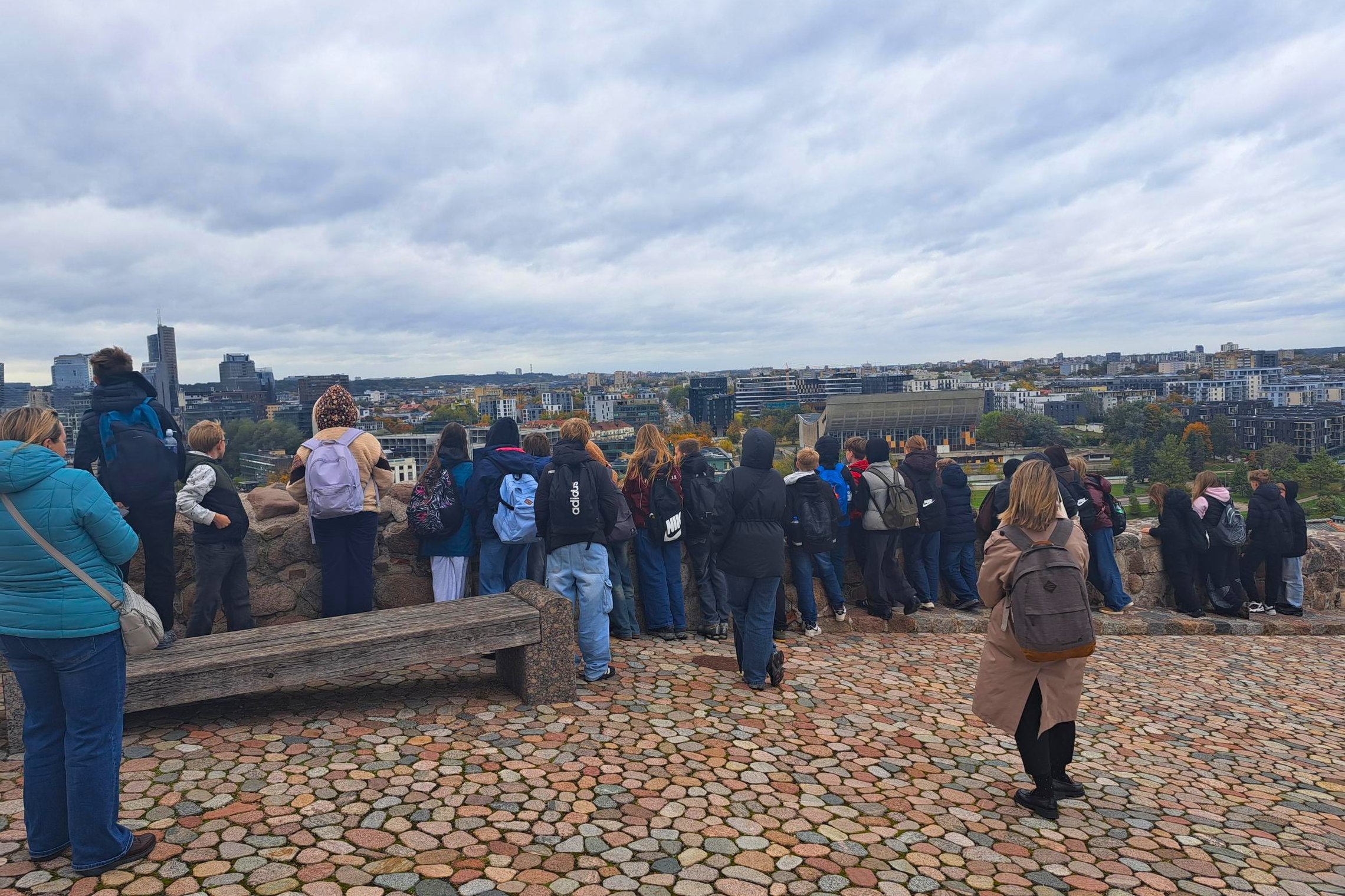 Klaipėdos Vydūno gimnazijos šeštokai, lydimi suaugusiųjų, nuo Vilniaus pilių teritorijoje esančios apžvalgos aikštelės grožisi sostinės panorama. Mokiniai stebi miesto pastatus ir rudenėjančius medžius, o virš Vilniaus driekiasi debesuotas dangus.