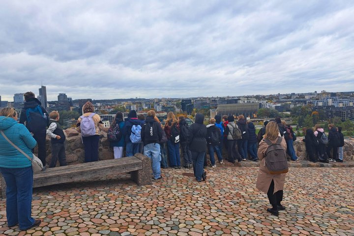 Klaipėdos Vydūno gimnazijos šeštokai, lydimi suaugusiųjų, nuo Vilniaus pilių teritorijoje esančios apžvalgos aikštelės grožisi sostinės panorama. Mokiniai stebi miesto pastatus ir rudenėjančius medžius, o virš Vilniaus driekiasi debesuotas dangus.