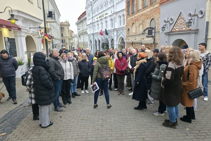 Ekskursijos dalyviai klausosi gidės pasakojimo Vilniaus senamiestyje, apsupti istorinių pastatų.