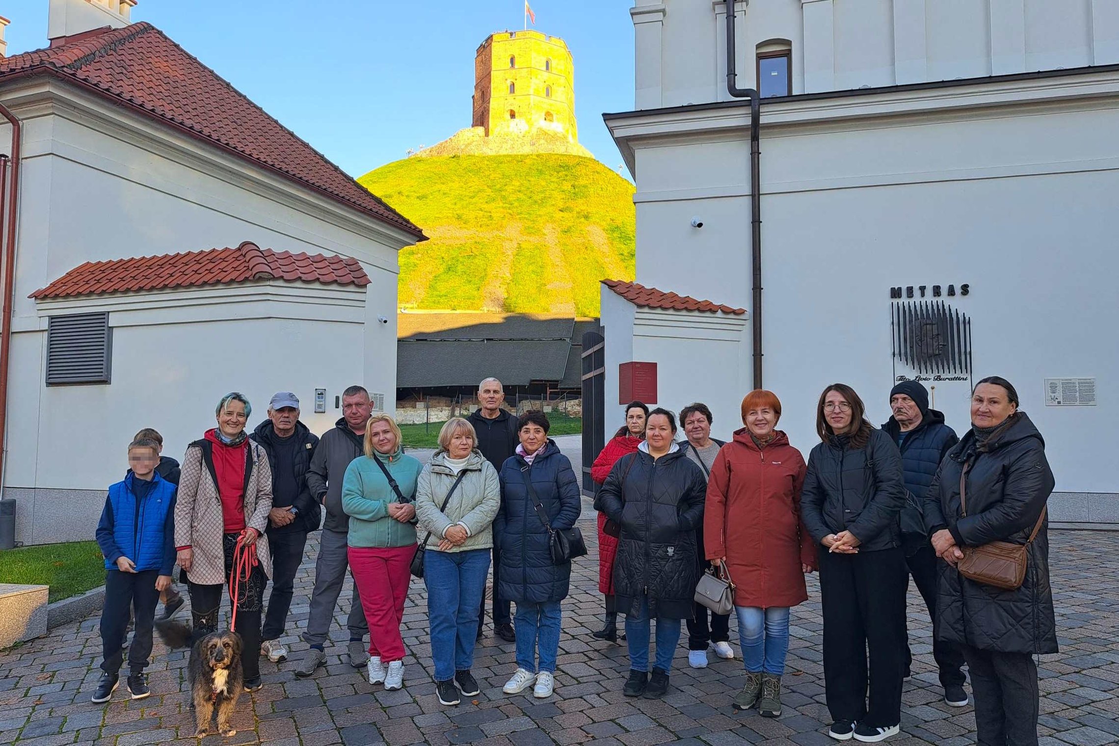 Ekskursijos „Valdovas spaudžia leteną: Vilniaus pilių šunys“ dalyviai su keturkoju palydovu fotografuojasi Vilniaus pilių teritorijoje. Fone matomas rudens saulės apšviestas Gedimino pilies bokštas ir istoriniai pastatai.