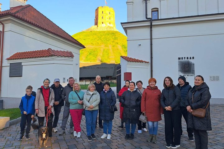 Ekskursijos „Valdovas spaudžia leteną: Vilniaus pilių šunys“ dalyviai su keturkoju palydovu fotografuojasi Vilniaus pilių teritorijoje. Fone matomas rudens saulės apšviestas Gedimino pilies bokštas ir istoriniai pastatai.