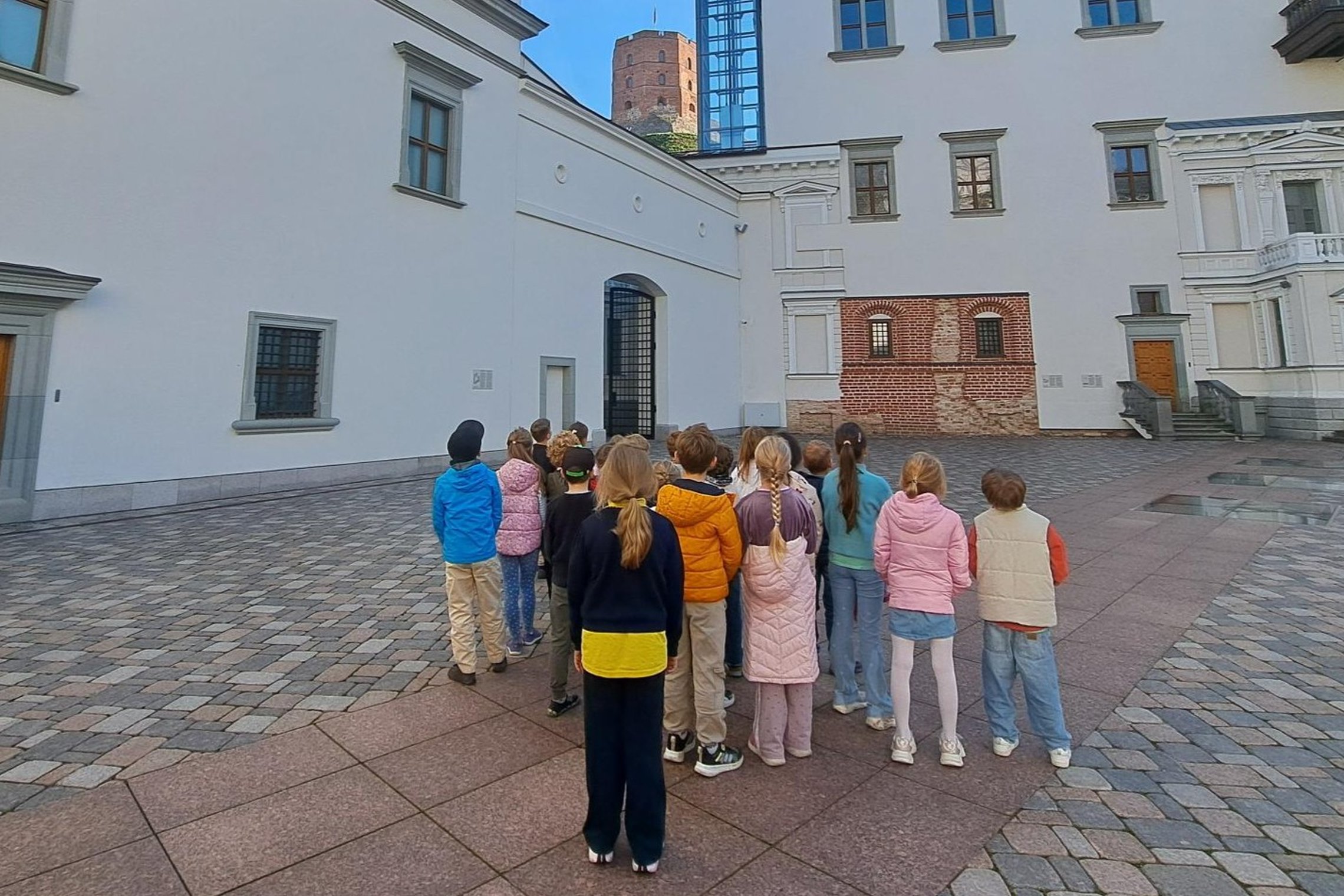 Nuotraukoje vaikai, Valdovų rūmų kieme, žiūri į Aukštutinės pilies bokštą. Jie dalyvauja edukacinėje pamokoje „Gediminas ir Vilniaus trys pilys“