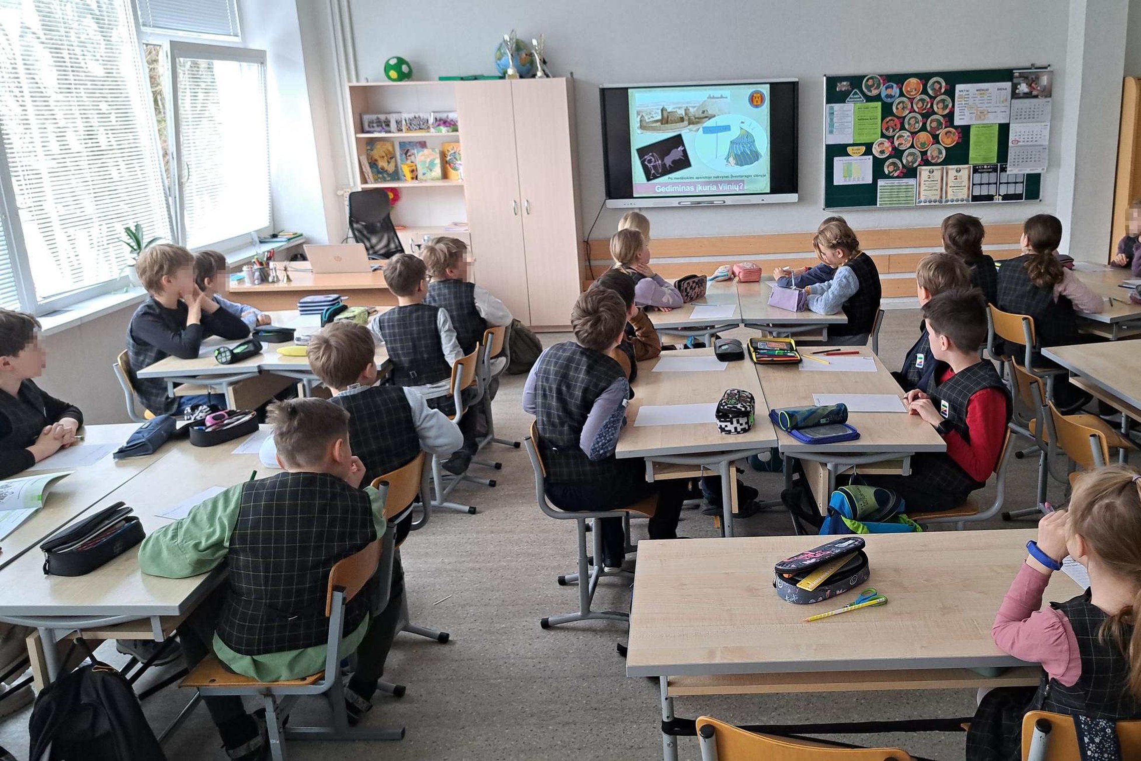 Trečiokai klasėje sėdi prie suolų ir stebi ekrane rodomą skaidrę apie Lietuvos Didžiuosius kunigaikščius; ant suolų padėtos pratybos ir penalai, priekyje – interaktyvus ekranas, lenta su mokinių darbais ir spintos su mokymo priemonėmis, pro langus sklinda