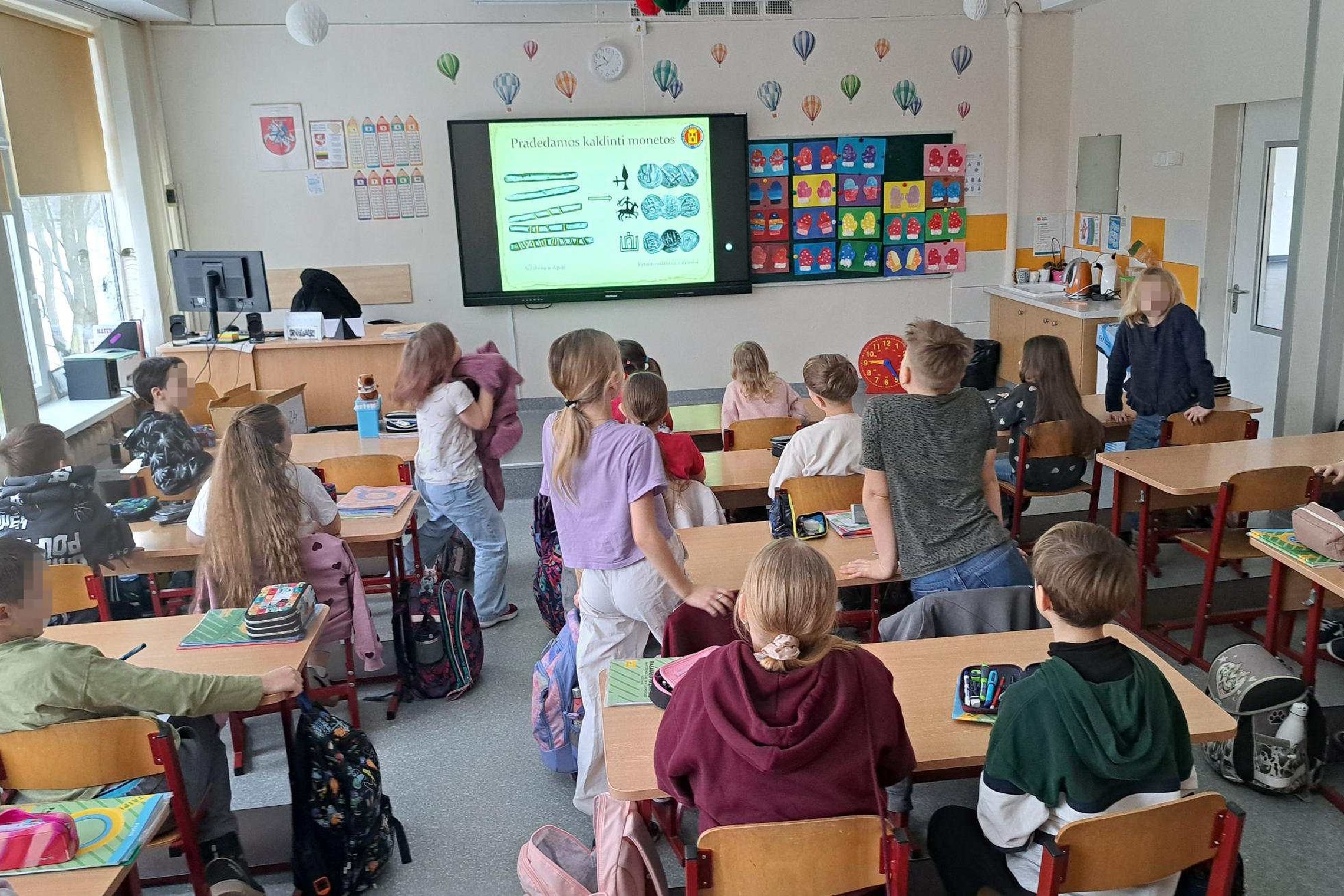 Trečiokai klasėje stebi edukacinę skaidrę apie monetų kaldinimą Lietuvos didžiųjų kunigaikščių laikais, sėdėdami suoluose ir aktyviai dalyvaudami istorijos pamokoje.