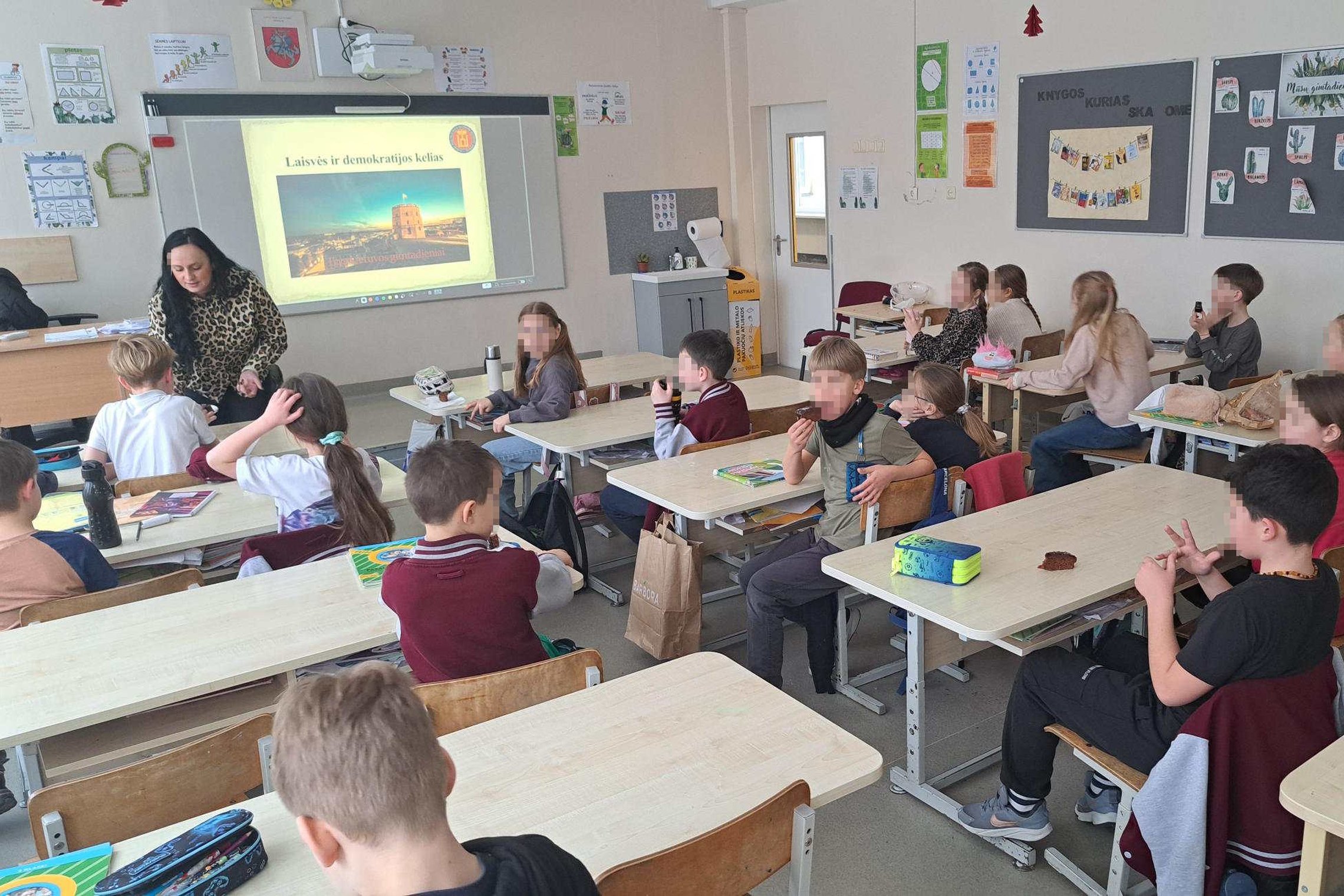 Trečiokai klasėje sėdi suoluose ir klausosi edukatorės, kuri prie interaktyvios lentos pristato temą „Laisvės ir demokratijos kelias“, rodomą su Gedimino pilies bokšto iliustracija.
