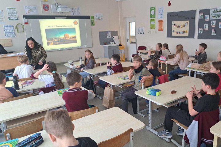 Trečiokai klasėje sėdi suoluose ir klausosi edukatorės, kuri prie interaktyvios lentos pristato temą „Laisvės ir demokratijos kelias“, rodomą su Gedimino pilies bokšto iliustracija.