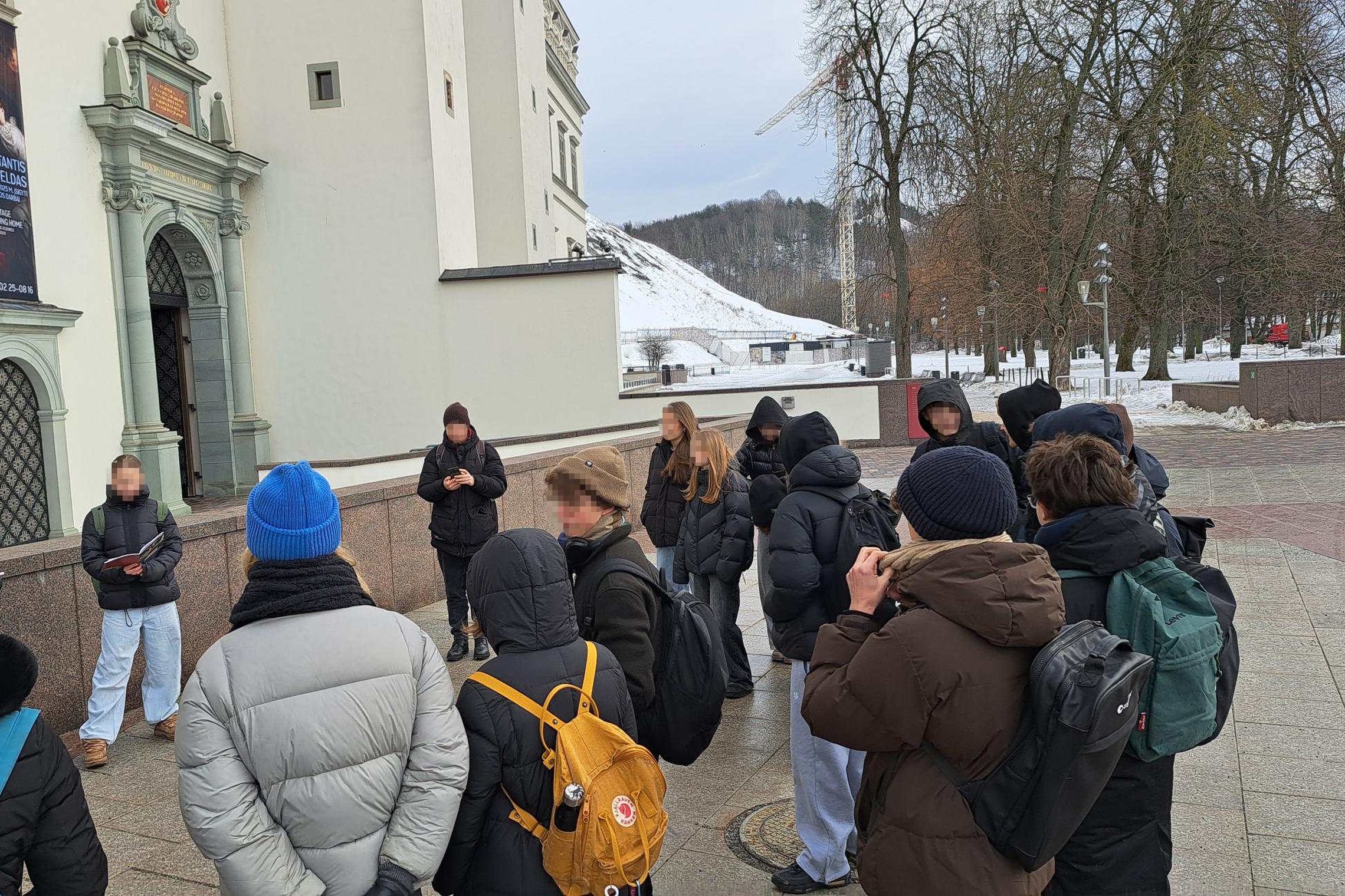 Lauke prie istorinio pastato susibūrę devintokai klausosi bendraklasio pasakojimo – mokiniai vilki žiemines striukes, stovi puslankiu prie įėjimo su dekoratyviniu portalu, fone matyti apsnigtas kalnas ir medžiai.
