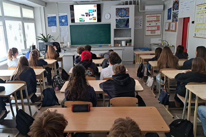Klasėje sėdintys aštuntokai klausosi edukacijos – mokiniai sėdi suoluose atsisukę į lentą ir ekraną, kuriame rodoma pristatymo skaidrė apie klimato kaitą; priekyje matyti mokytojo darbo vieta, lenta, plakatų stendas ir kriauklė, pro langus į klasę patenk
