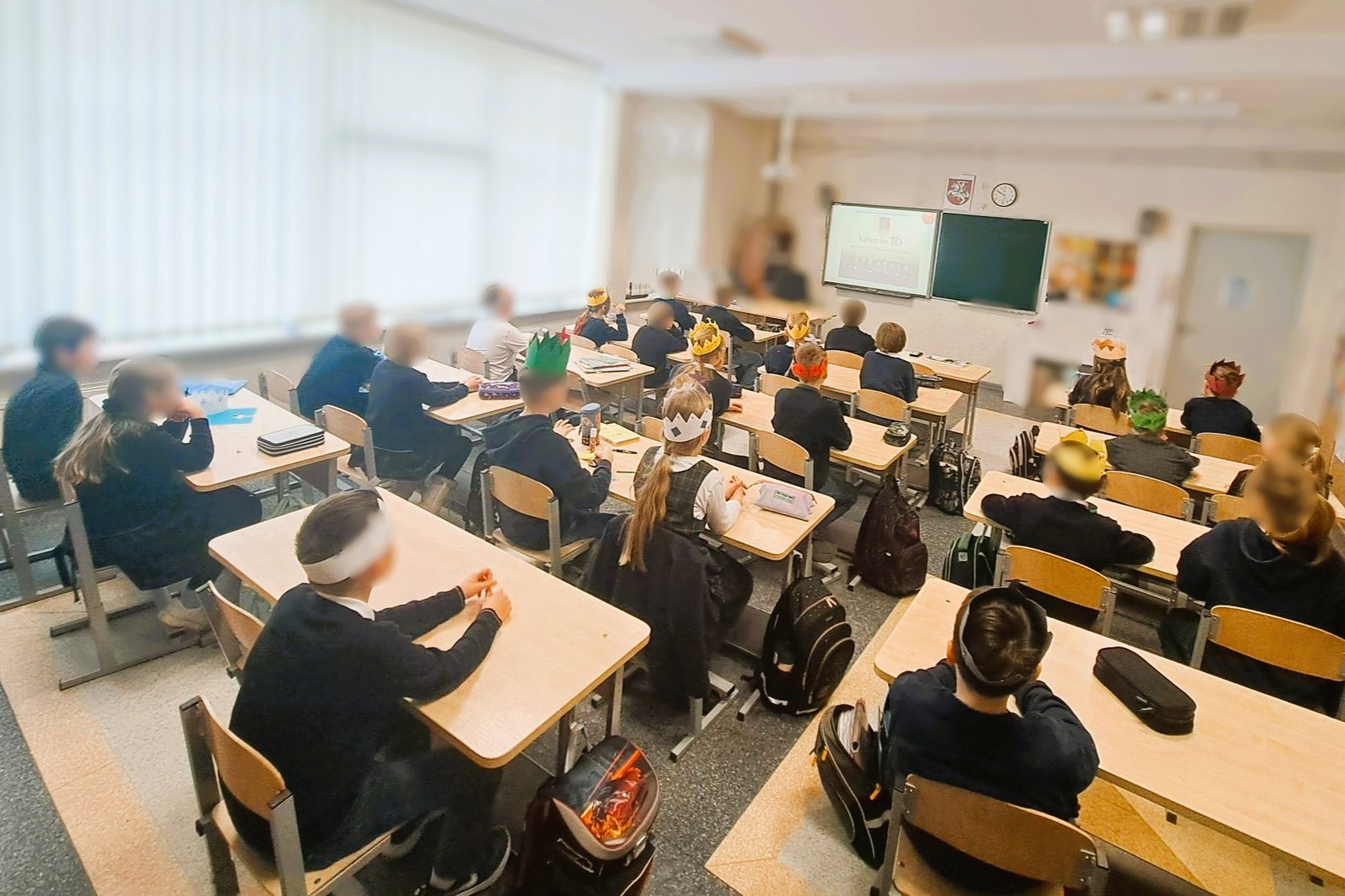 Nuotraukoje klasės vaizdas ir joje esantys mokiniai. Jie sėdi suoluose ir žiūri į ekraną. Ekrane edukacijos vaizdinė medžiaga.