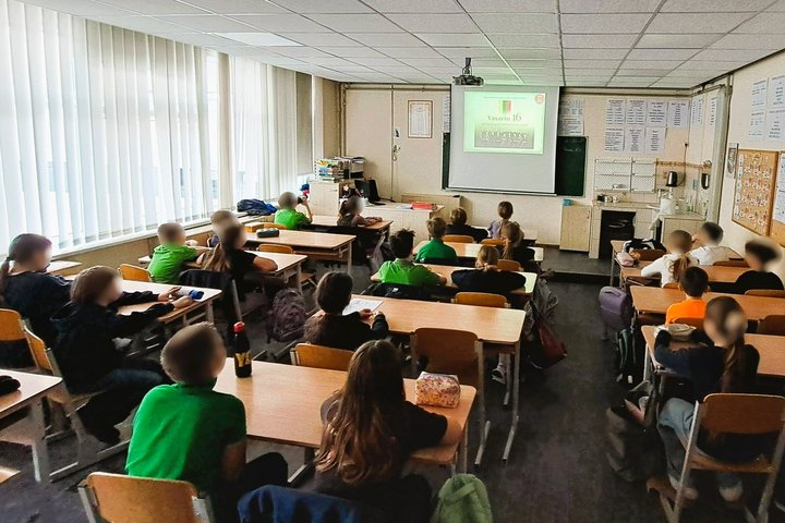 Nuotraukoje mokiniai klasėje. Sėdi suoluose ir žiūri į ekraną. Ekrane Vasario 16-osios Akto signatarai