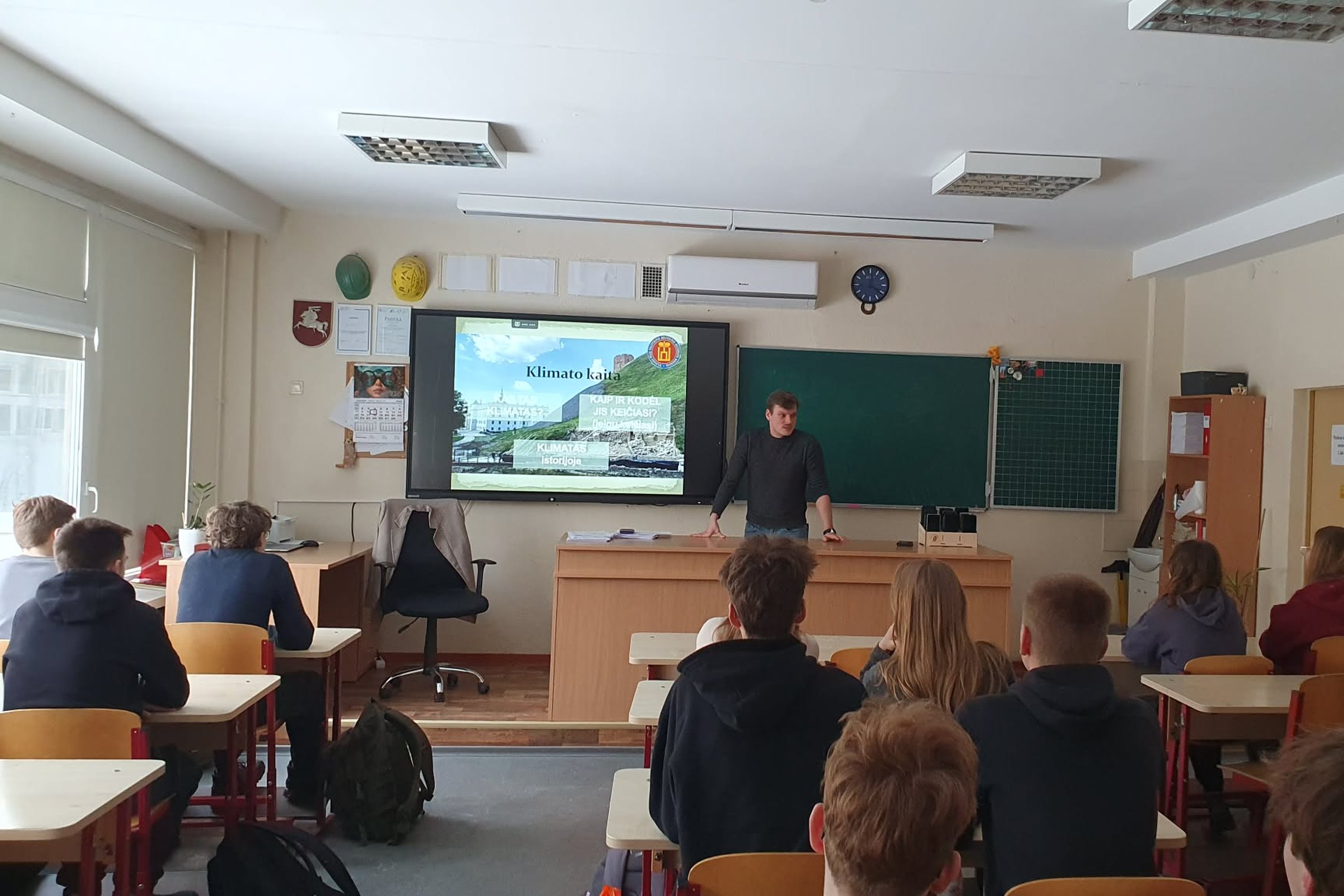Edukatorius stovi klasės priekyje prie interaktyvios lentos su pristatymu „Klimato kaita“, o suoluose sėdintys mokiniai klausosi edukacijos apie klimato ir gamtos pokyčius.
