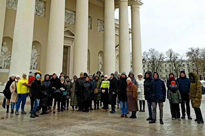 Grupė žmonių žiemos metu stovi Vilniaus katedros aikštėje priešais Katedros kolonadą; visi apsirengę šiltais paltai­s ir kepurėmis, ekskursijos dalyviai fotografuojasi po atviru dangumi.