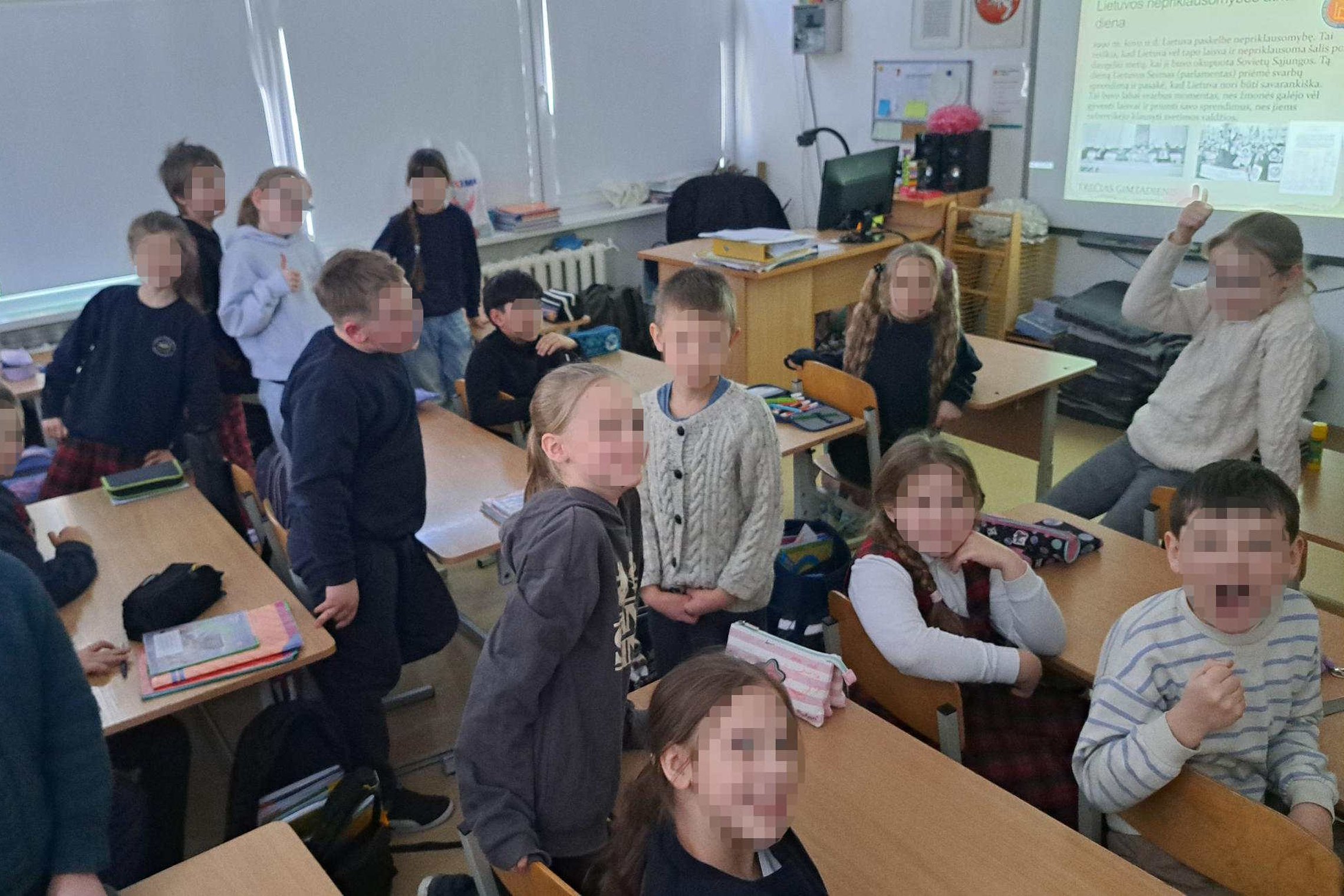 Trečiokų klasė edukacijos metu: mokiniai stovi ir sėdi suoluose, klasėje matomas mokytojo stalas ir ekrane rodomas pristatymas apie Lietuvos nepriklausomybę.