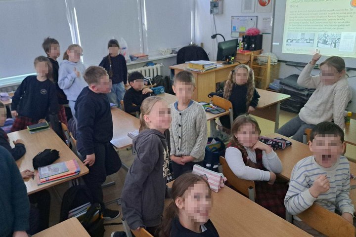 Trečiokų klasė edukacijos metu: mokiniai stovi ir sėdi suoluose, klasėje matomas mokytojo stalas ir ekrane rodomas pristatymas apie Lietuvos nepriklausomybę.