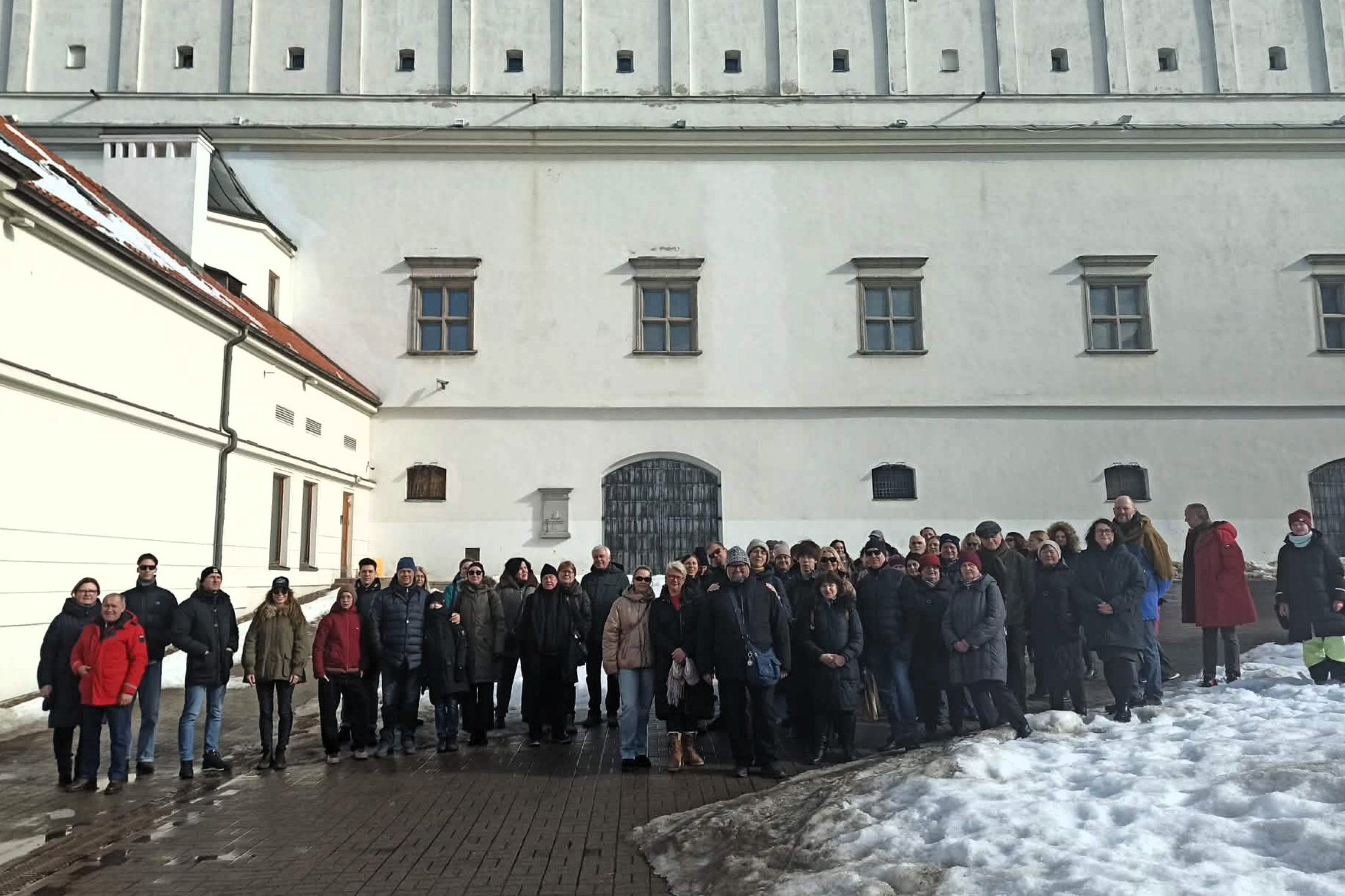 Ekskursijos dalyvių grupė stovi Vilniaus pilių teritorijoje prie balto mūrinio pastato sienos; šalia matyti tirpstančio sniego krūva ir saulėta, bet vėsi žiemos diena.
