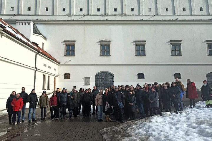 Ekskursijos dalyvių grupė stovi Vilniaus pilių teritorijoje prie balto mūrinio pastato sienos; šalia matyti tirpstančio sniego krūva ir saulėta, bet vėsi žiemos diena.