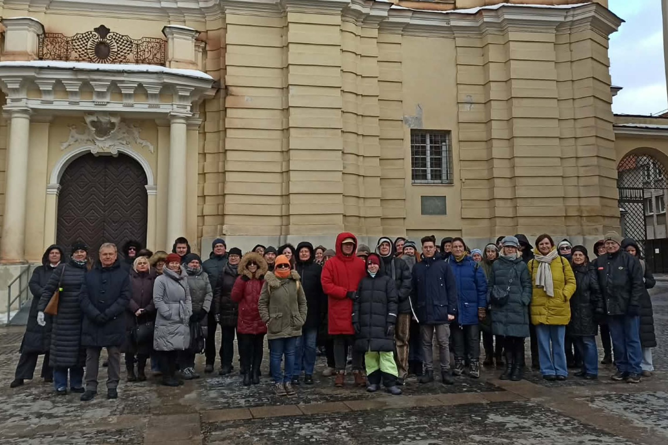 Ekskursijos dalyvių grupė stovi prie barokinės Šv. Jonų bažnyčios, prie puošnaus barokinio įėjimo su kolonomis; žmonės apsirengę žieminiais paltais, matyti drėgna, šalta diena.