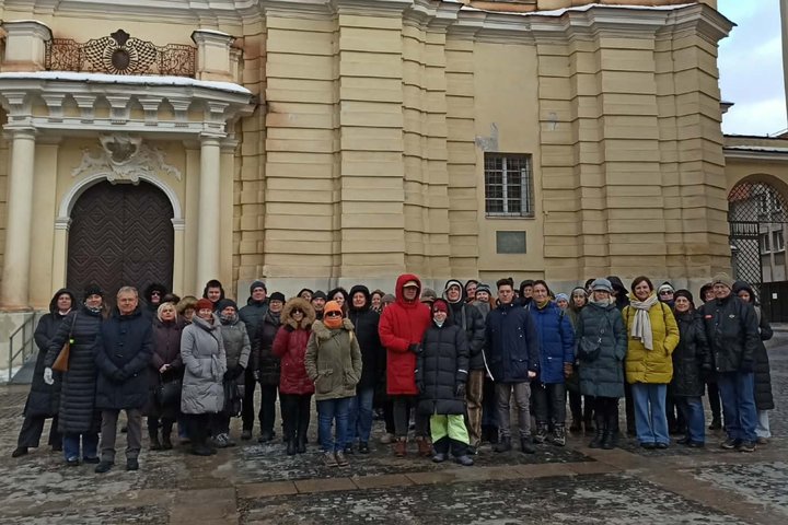 Ekskursijos dalyvių grupė stovi prie barokinės Šv. Jonų bažnyčios, prie puošnaus barokinio įėjimo su kolonomis; žmonės apsirengę žieminiais paltais, matyti drėgna, šalta diena.