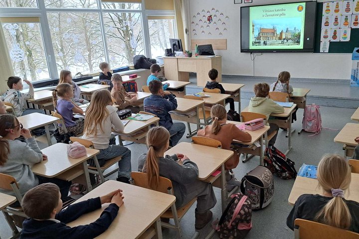 Klasėje sėdintys trečios klasės mokiniai klausosi edukacijos – priekyje ekrane rodoma skaidrė apie Vilniaus katedrą ir Žemutinę pilį, ant suolų – sąsiuviniai ir kuprinės, pro langus matyti žiemos vaizdas.