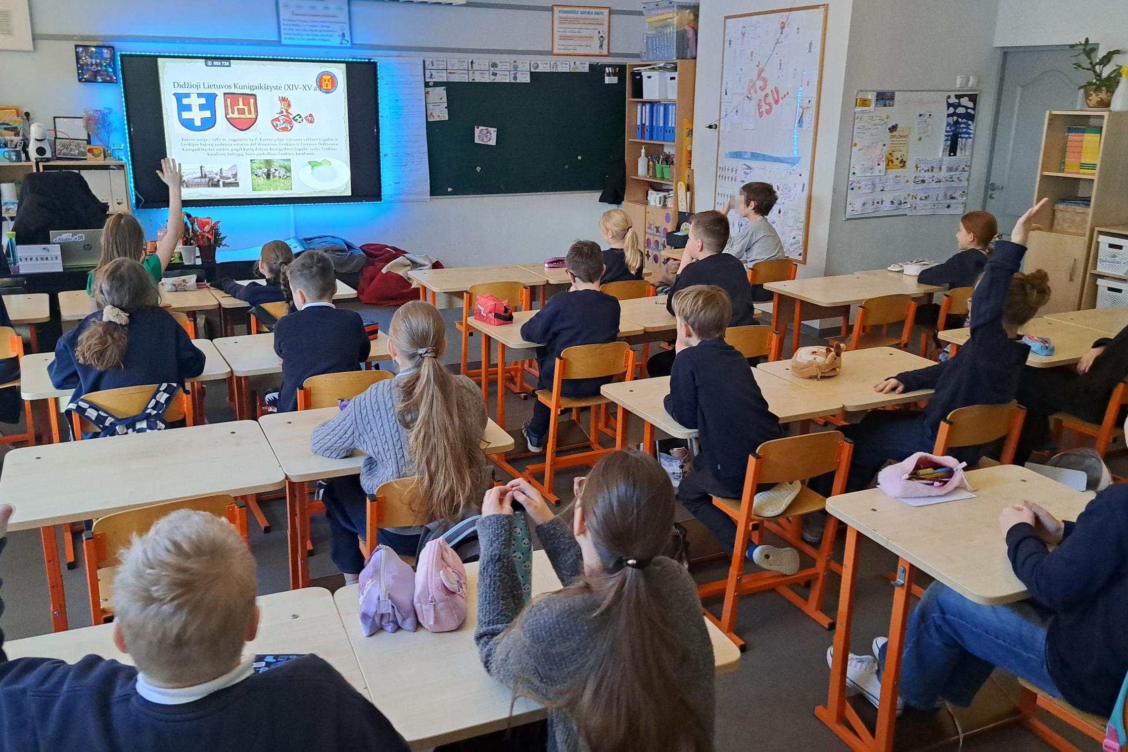 Ketvirtokai sėdi klasėje ir pakeltomis rankomis dalyvauja edukacijoje apie Lietuvos laisvės ir demokratijos kelią, ekrane rodoma prezentacija apie Didžiąją Lietuvos Kunigaikštystę ir Vilniaus pilis.