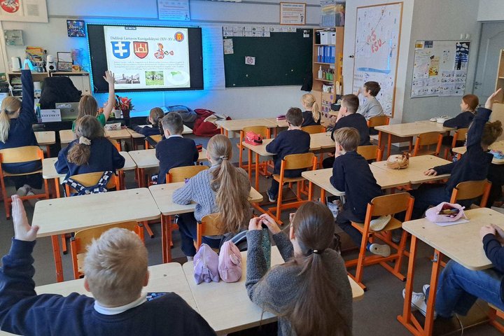 Ketvirtokai sėdi klasėje ir pakeltomis rankomis dalyvauja edukacijoje apie Lietuvos laisvės ir demokratijos kelią, ekrane rodoma prezentacija apie Didžiąją Lietuvos Kunigaikštystę ir Vilniaus pilis.