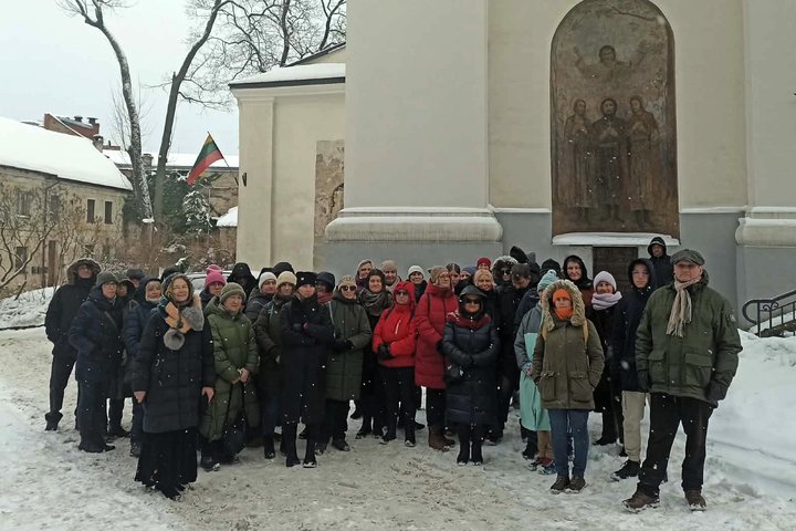 Didelė žmonių grupė žieminiais drabužiais stovi lauke ant sniego prie bažnyčios fasado su religine freska. Fone matyti pastatai, medžiai be lapų ir plevėsuojanti Lietuvos vėliava. Sninga.