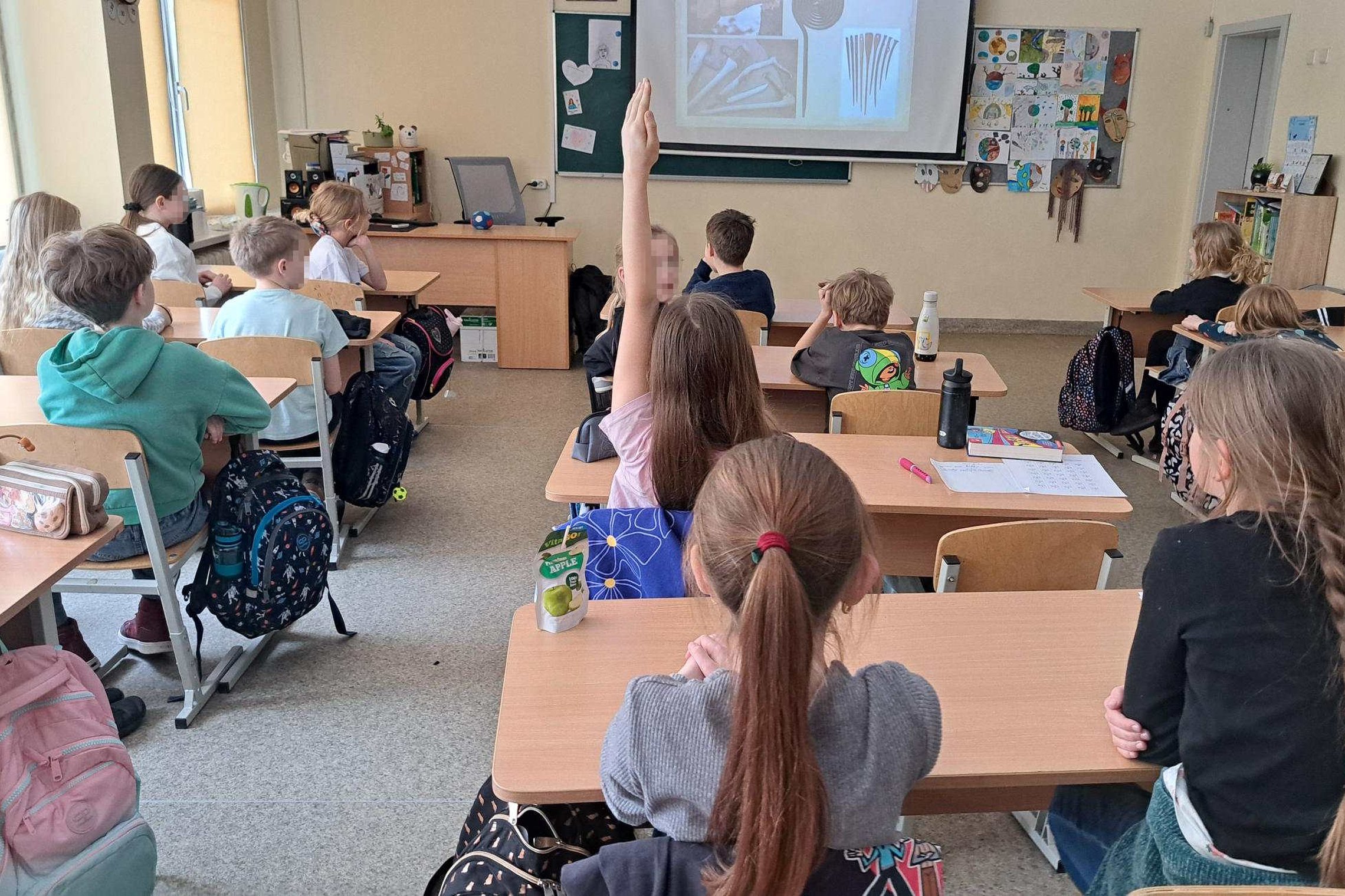 Klasėje sėdintys trečiokai klausosi edukacijos – mokiniai sėdi suoluose atsisukę į ekraną su rodomomis skaidrėmis, viena mokinė pakėlusi ranką atsakyti, ant sienų matyti vaikų piešiniai ir mokymo priemonės.