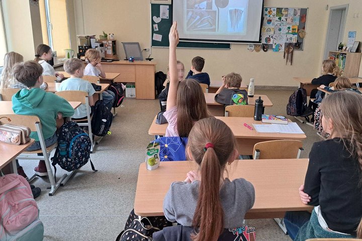 Klasėje sėdintys trečiokai klausosi edukacijos – mokiniai sėdi suoluose atsisukę į ekraną su rodomomis skaidrėmis, viena mokinė pakėlusi ranką atsakyti, ant sienų matyti vaikų piešiniai ir mokymo priemonės.