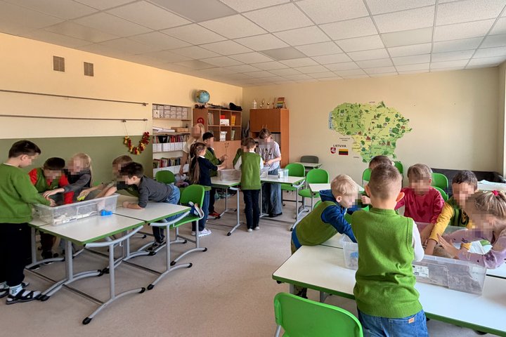 Vaikai klausosi edukatorės pasakojimo, rankose mokslinės priemonės; fone matyti žalias Lietuvos žemėlapis.