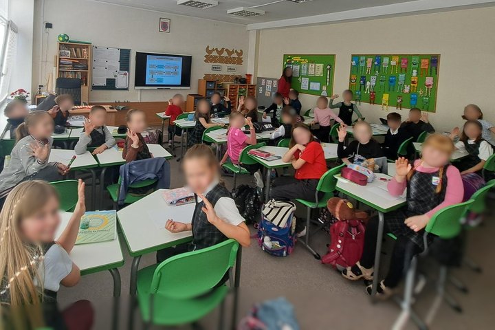 Nuotraukoje mokiniai klasėje. Jie sėdi suoluose. Mojuoja. Vaikai dalyvauja edukacijoje.